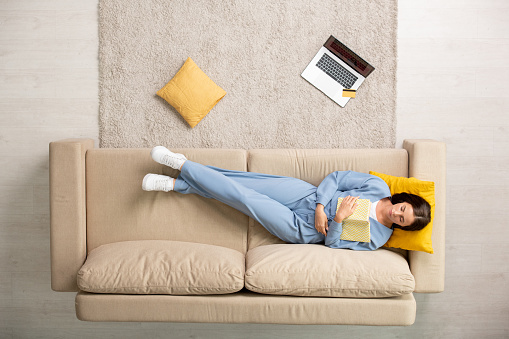 Top view of restful female in blue pajamas napping with open book on chest while lying on couch with head on yellow pillow after work