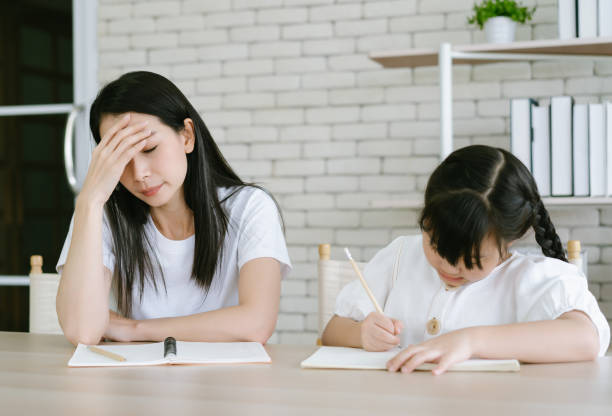 動揺アジアの母親は、小さな娘が教え、宿題の書き込みや読書をすることに怒っています。彼女は家の机の上に座っている間理解していない。子どもの教育問題、親子の対立。 - daughter distraught little girls worried ストックフォトと画像