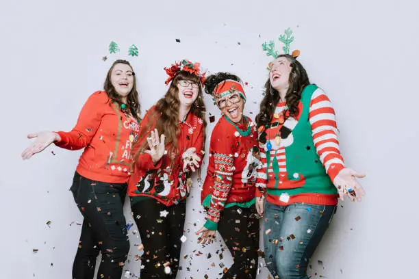 A group of adult women friends celebrate the holiday taking a portrait wearing "ugly" Christmas sweaters, throwing confetti into the air.