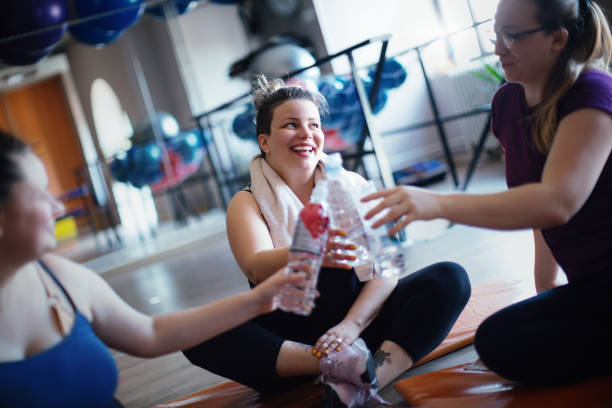 We managed to endure the workout to the end. Overweight women completed their workout in the gym. They are sitting on the floor and toasting with a water. women satisfaction decisions cheerful stock pictures, royalty-free photos & images