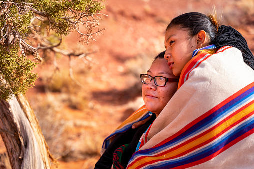 Two Native American, Navajo Sisters Dressed In Traditional Clothing, Wrapped In A Navajo Blanket Sitting Close Together, Hugging