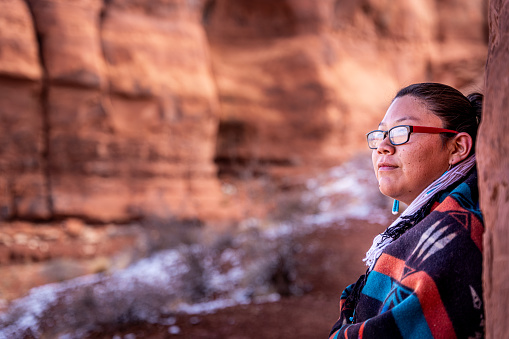 A Closeup Of A Confident Native American, Navajo Young Woman Wrapped Up In A Traditional Navajo Wool Blanket, Sitting In The Iconic Teardrop Arch, Monument Valley, Utah, Arizona