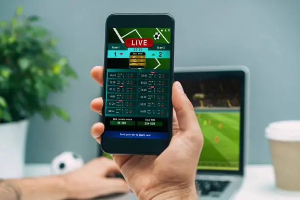 Close up cropped shot of male handholding mobile phone with bookmaker application interface. Man watching football play online broadcast on his laptop waiting for winning results.