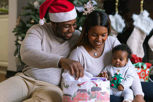 A young mixed race family open presents with their newborn daughter on Christmas Day. The baby is wearing a Christmas sweater. The dad is wearing a red Christmas hat.