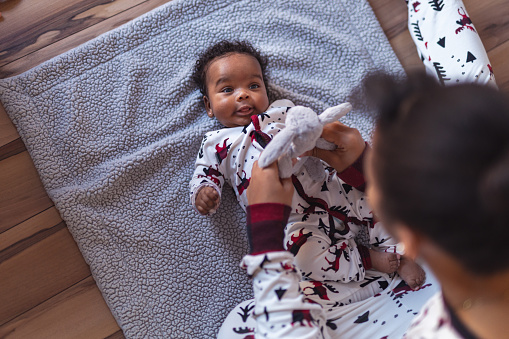 A sister of elementary school age is playing with her baby sister on Christmas Day. The baby girl is wearing a Christmas onesie and smiling up at her sister. Her sister is playfully holding a stuffed bunny over her face.