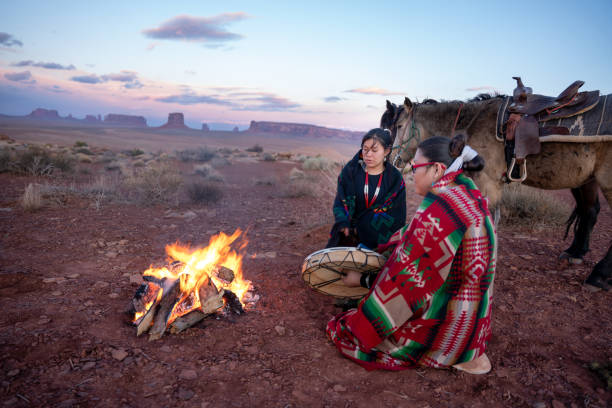 Photo of Two Native American Sisters, One Playing A Drum, Wrapped In Traditional Navajo Blankets Staying Warm By The Campfire Horses Behind Them, Iconic Monuments, Sunset