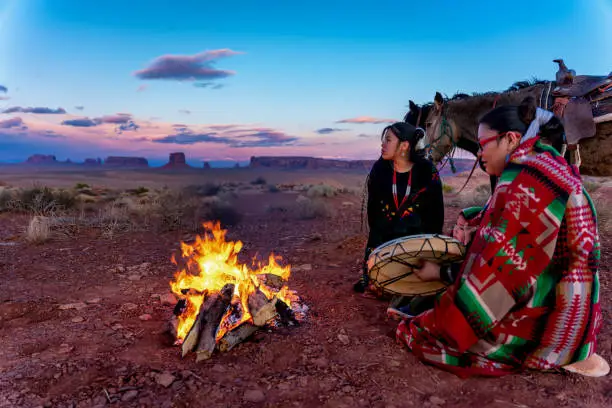 Photo of Two Native American Sisters And Their Little Brother Wrapped In Traditional Navajo Blankets Staying Warm By The Campfire, Their Horses Behind The Iconic Monuments And Sunset In The Background