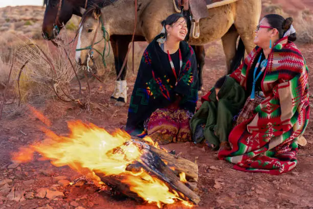 Photo of Two Native American Sisters And Their Little Brother Wrapped In Traditional Navajo Blankets Staying Warm By The Campfire, Their Horses Behind The Iconic Monuments And Sunset In The Background