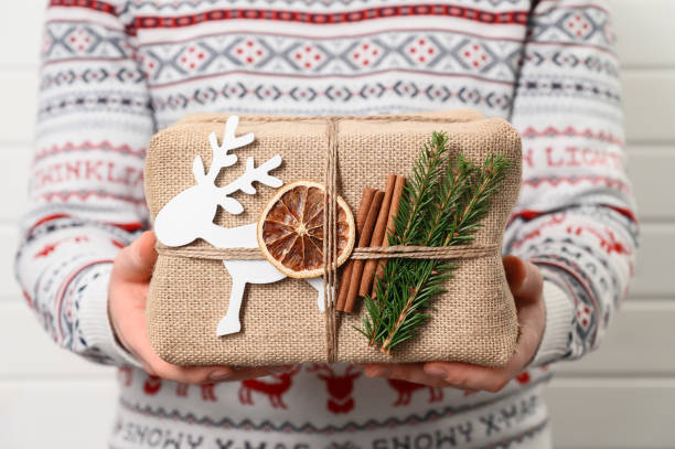 delivery man holding decorated gift box in eco-friendly reusable packaging. man in sweater holds christmas present - gift orange green package imagens e fotografias de stock