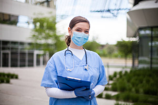 Doctor in front of hospital Female doctor holding blue clipboard standing outside hospital or clinic, frontline key medical worker portrait in modern care facility or nursing home complex yard,Coronavirus pandemic outbreak crisis frontline worker mask stock pictures, royalty-free photos & images