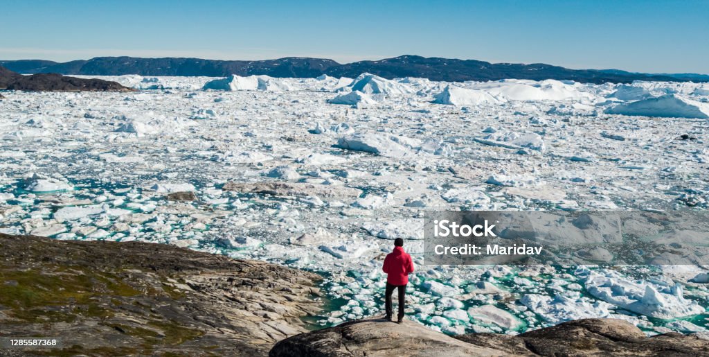 Travel in arctic landscape nature with icebergs - Greenland tourist man explorer Travel in arctic landscape nature with icebergs - Greenland tourist man explorer - tourist person looking at amazing view of Greenland icefjord - aerial drone image. Man by ice and iceberg, Ilulissat. Greenland Stock Photo