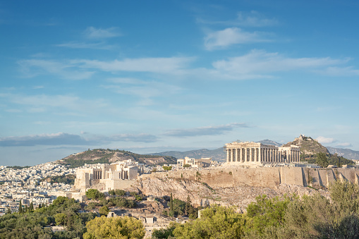 Bask in the brilliance of a sunny summer day as you behold the stately main building of the Academy of Athens, a jewel within Theophil Hansen's esteemed Trilogy, situated in central Athens, Greece. Against a backdrop of azure skies, this neoclassical masterpiece radiates timeless elegance and scholarly reverence. Its majestic columns and intricate architectural details stand in harmonious contrast to the vibrant greenery that surrounds it. As sunlight bathes the scene in a golden glow, the building invites contemplation and reflection, embodying the pursuit of knowledge and intellectual enlightenment