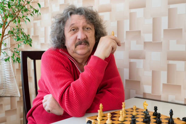 a 70-year old man with curly hair sitting at the table and plain chess. - 60 70 year old imagens e fotografias de stock