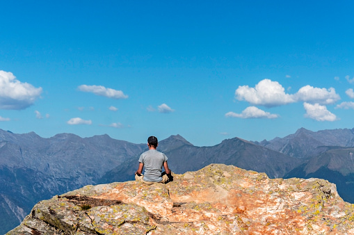 young man from back meditating on rock against background of Caucasus mountains and blue cloudy sky. Relaxation in nature, solo travel, hike in mountains, landscape