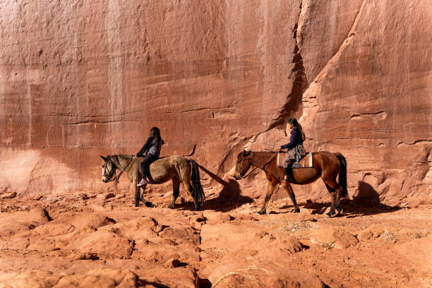 ein junger bruder und schwester reiten ihre pferde durch eine massive felswand gefüllt mit nativen petroglyphen in monument valley, arizona - monument valley navajo mesa monument valley tribal park stock-fotos und bilder