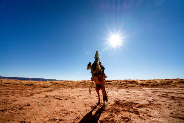 ein junger navajo junge reitet sein pferd durch die wüste in der nähe von camel butte, mit monument valley im hintergrund - monument valley navajo mesa monument valley tribal park stock-fotos und bilder