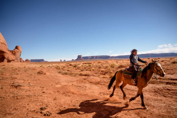 ein junger navajo junge reitet sein pferd durch die wüste in der nähe von camel butte, mit monument valley im hintergrund - monument valley navajo mesa monument valley tribal park stock-fotos und bilder