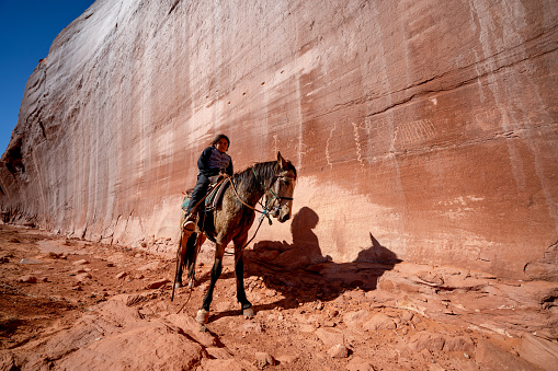 A Young Native American, Navajo Boy Rides His Horse Near A Massive Rock Wall Filled With Native Petroglyphs