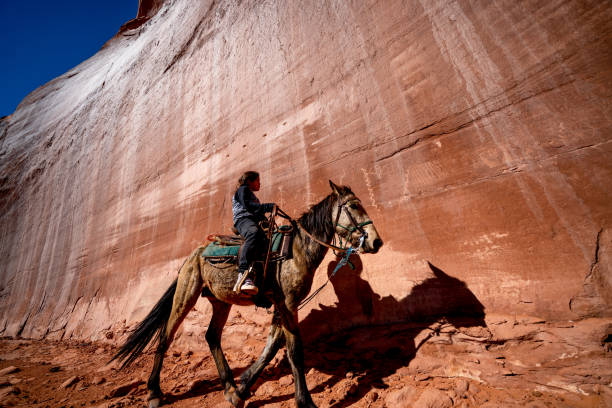 ein junger indianer, navajo boy reitet sein pferd in der nähe einer massiven felswand gefüllt mit native petroglyphen - monument valley navajo mesa monument valley tribal park stock-fotos und bilder