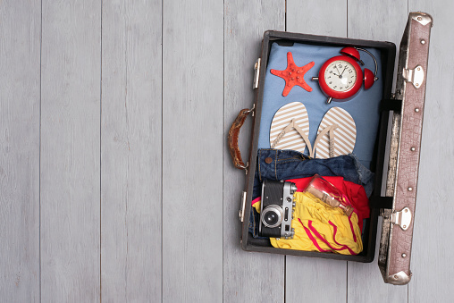 Open suitcase with clothing and photo camera on the wooden flat lay floor background with copy space.