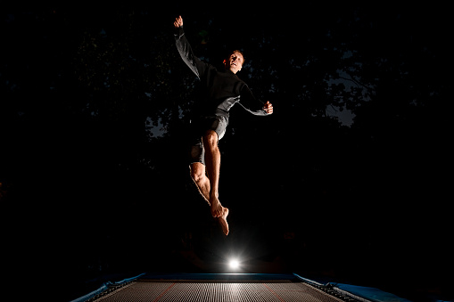 cheerful young man in black clothes jumps in the air on trampoline at dark time