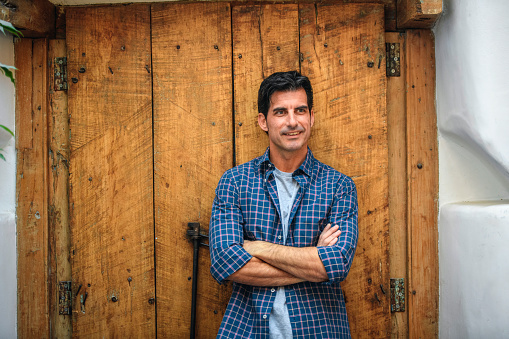 Relaxed man in early 40s wearing casual clothing and looking away from camera while standing against rustic wood background with arms crossed.