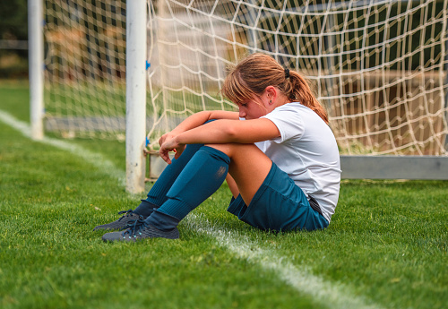 Side view of tired 9 year old female footballer sitting on field next to goal thinking about how she played in practice.