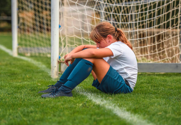 Young Girl Footballer Sitting on Field After Practice Side view of tired 9 year old female footballer sitting on field next to goal thinking about how she played in practice. disappointment stock pictures, royalty-free photos & images