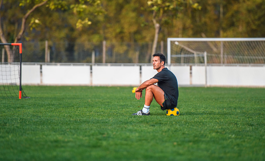 Fit male footballer in early 40s wearing black shirt and shorts and sitting on ball in middle of sports field after practice.