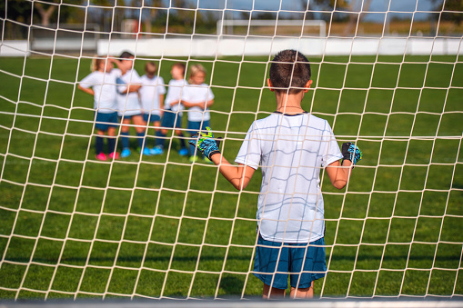 Rear view through netting of young boy footballer with gloved hands up ready to defend against practice kicks.