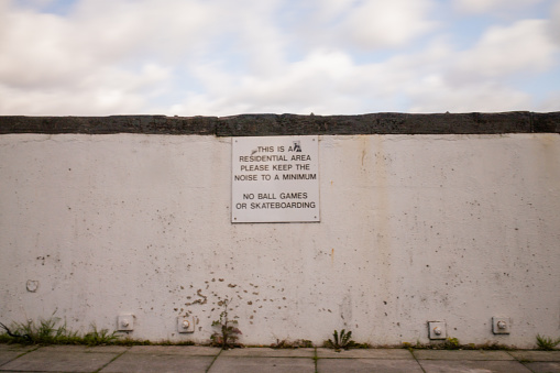 Picture of a white wall with a white sign requesting to keep the noise to a minimum, and prohibiting skateboarding and ball games, and with a slightly cloudy sky as background