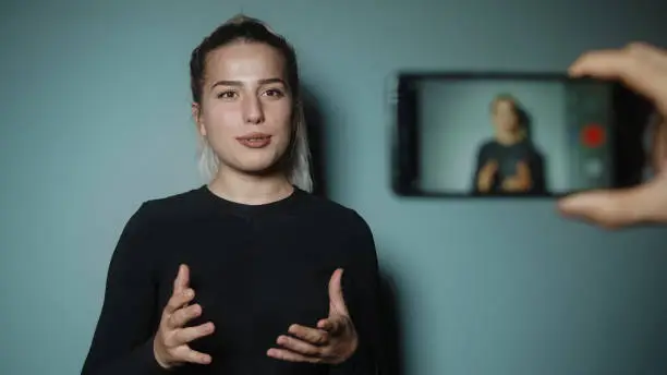 Beautiful smiling fashion blogger blonde woman making online new video blog, vlog, with her smartphone on studio background. Influencer speaking to smartphone during conference call record