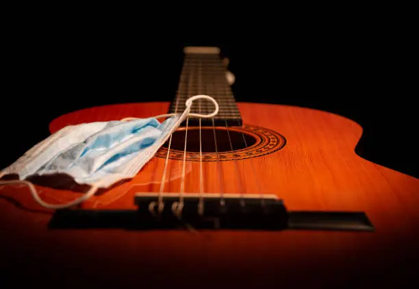 This studio shot is a low shot of the main body and neck of an acoustic guitar. Shot with a warm, moody light highlighting a face covering mask. A concept for covid-19 coronavirus laws and restrictions for performing live musicians, where face coverings are a requirement for gigging stage performance and public attendance.