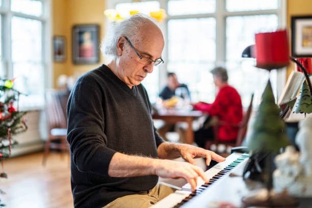 reunión familiar navideña. el hombre mayor de 72 años de edad de pelo plateado, el músico, tocando el piano para su familia que está cenando en el telón de fondo en el amplio y luminoso salón decorado para navidad. - senior adult 60 65 years people occupation fotografías e imágenes de stock