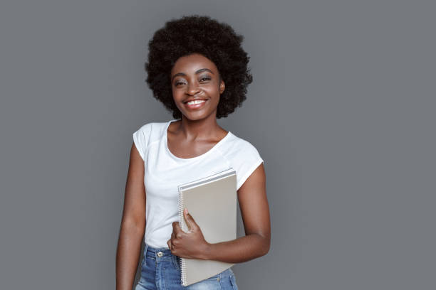 istock Young woman standing isolated on gray with notebook looking camera happy 1285703077