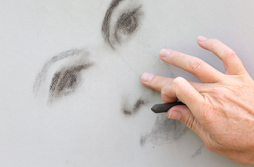 One hand draws a woman's face in brown pastel chalk on gray textured paper