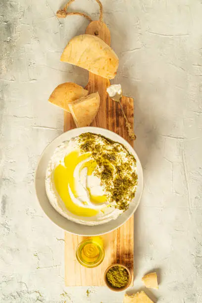 Photo of Popular middle eastern appetizer labneh or labaneh, soft white goat milk cheese with olive oil, hyssop or zaatar, served with pita bread on a wooden plate on a grey table,