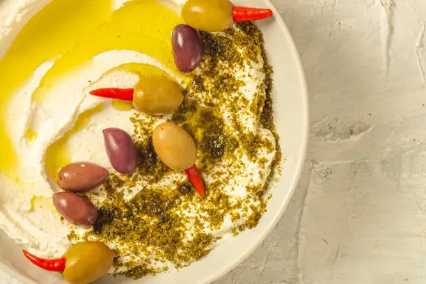 Photo of Popular middle eastern appetizer labneh or labaneh, soft white goat milk cheese with olive oil, hyssop or zaatar, olives on grey table, top view ,flat lay. Close up image.