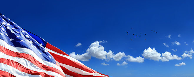 Close up waving American flag at tall flagpole over clear blue sky with white clouds.