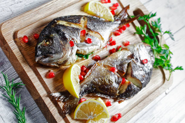 Whole baked dorado fish stuffed with fresh parsley on a cutting board with lemon wedges and pomegranate seeds on white wooden table, close-up, top view
