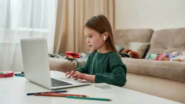 A little girl enjoying her time with a laptop being in a huge bright guestroom sitting at a table with her wireless earphones on during distance education alone. Homeschooling concept