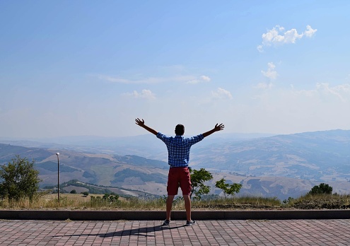 Young man is standing on a cobblestone road with trees and mountains in the distance with his arms outstretched to his sides