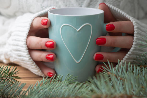 Woman's hands in white knitted sweater with red nail polish hold a mint mug with heart near branches of Christmas tree. A woman's hands in a white knitted sweater with red nail polish hold a mint mug with a heart, near the branches of a Christmas tree. christmas nails stock pictures, royalty-free photos & images