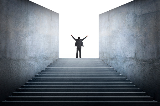A businessman stands with his arms raised at the top of a long and wide set of steps surrounded by tall concrete walls.