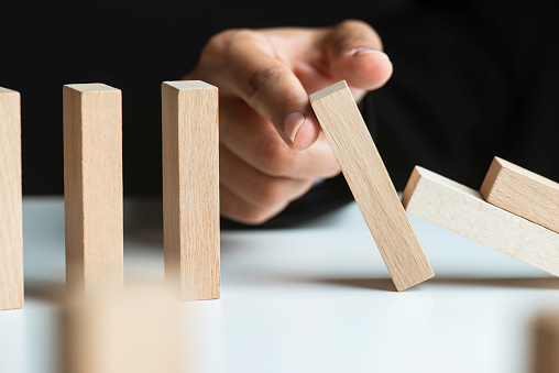 Finger is stopping chain reaction of dominos who are about to fall in front of a dark background.