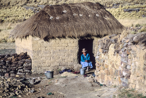 Peruvian Plateau, Peru - Aug 1994: A woman in traditional Peruvian clothing enjoys the sun sitting on the threshold of her house built with dried mud bricks and a thatched roof.