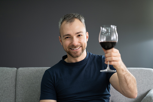 A portrait of an elegant handsome male sommelier visually inspecting a glass of wine in a wine cellar. It stands next to a row of wooden barrels. Professional degustation expert in winemaking.