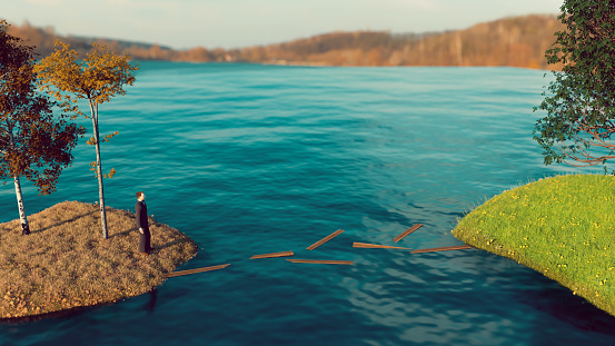 Man stands on island among dead leaves and looks at another island with green grass. There is water between the two islands. In the water is several planks from a bridge, that has been destroyed. Concept of showing the grass is always greener on the other side.