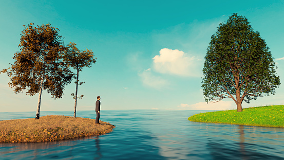 Man stands on island among dead leaves and looks at another island with green grass. There is water between the two islands. Concept of showing the grass is always greener on the other side.