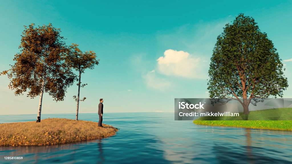 Concept of the grass is always greener on the other side Man stands on island among dead leaves and looks at another island with green grass. There is water between the two islands. Concept of showing the grass is always greener on the other side. The Grass Is Always Greener Stock Photo
